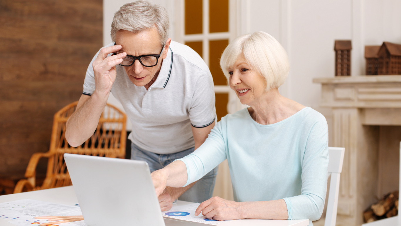 A senior woman points to her laptop while her husband peers over to look at it