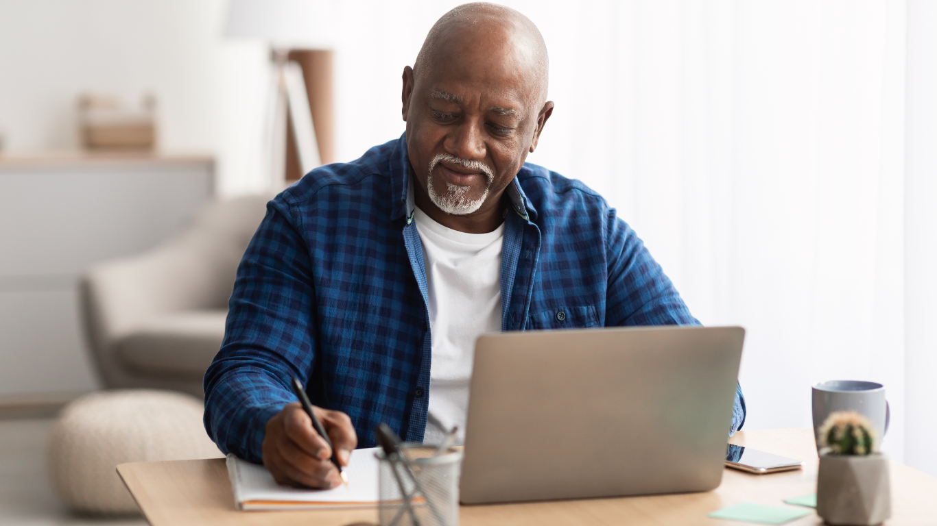 A senior man sits at a table at home and looks at his laptop and writes notes in a notebook